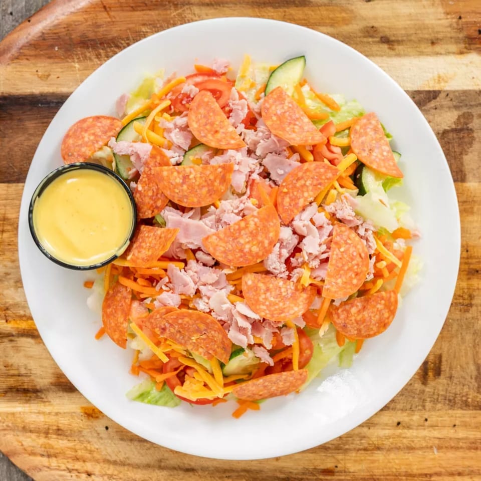 A vibrant chef salad with pepperoni, ham, cheese, and vegetables, served with a side of dressing.