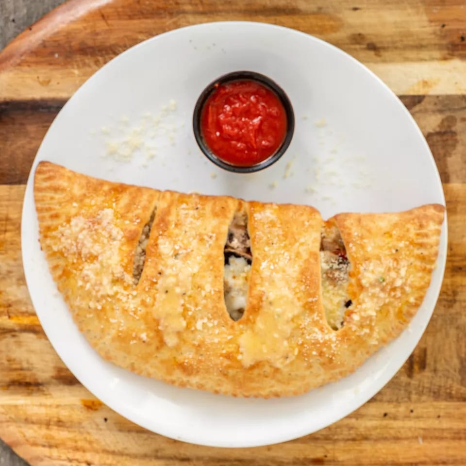 Golden-brown calzone with parmesan cheese and a side of red marinara sauce on a white plate.
