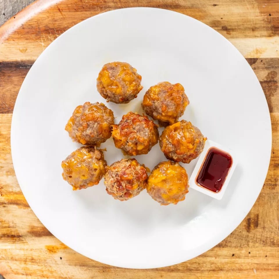 Eight glazed meatballs with melted cheese and a side of dipping sauce on a white plate.