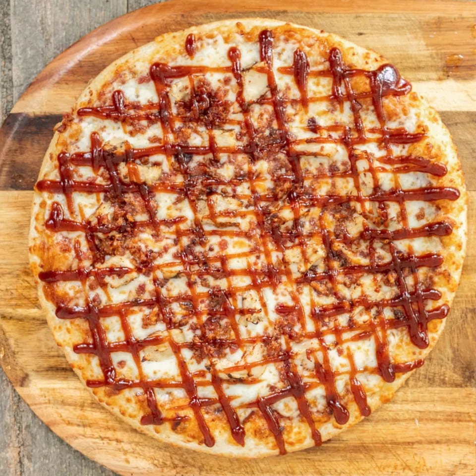 A BBQ chicken pizza with a lattice of sauce and bacon bits on a wooden board.