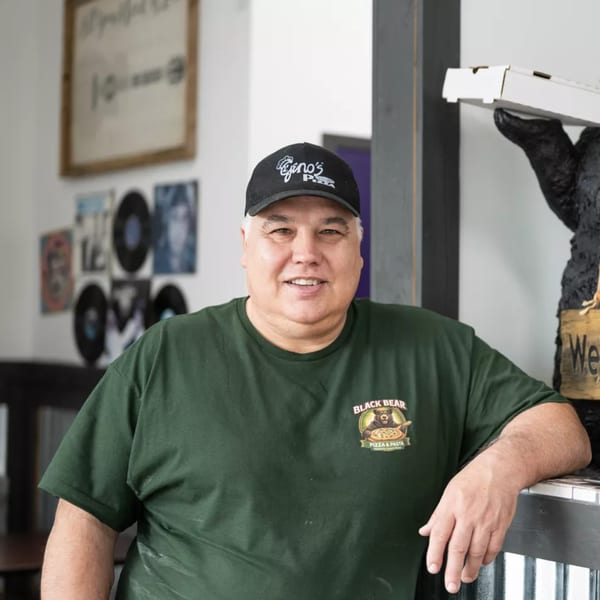 Man in a green Black Bear Pizza & Pasta shirt and Gino's Pizza hat smiles at the camera.